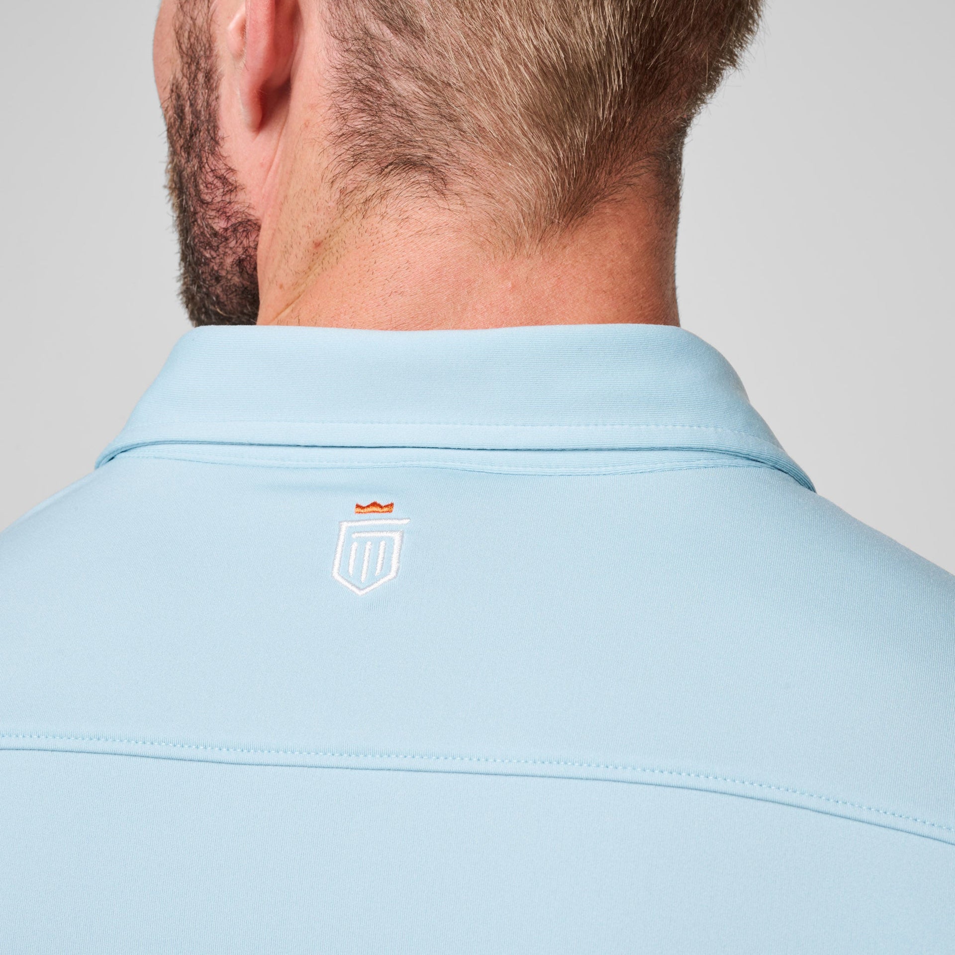 Light blue polo shirt with collar and small embroidered shield logo on back, shown on man from behind.