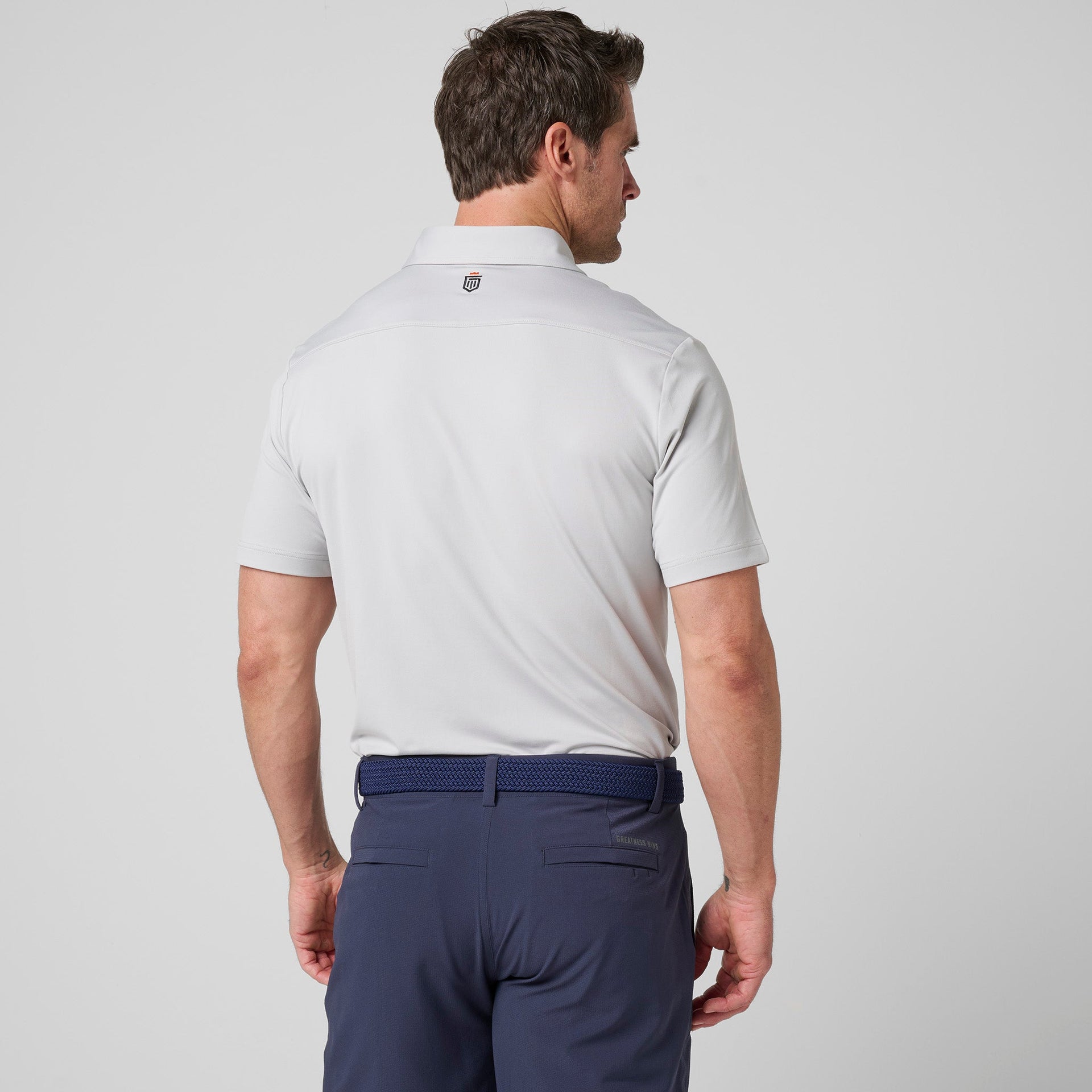 Man wearing white short-sleeve polo shirt with small logo on back collar and navy pants with belt.