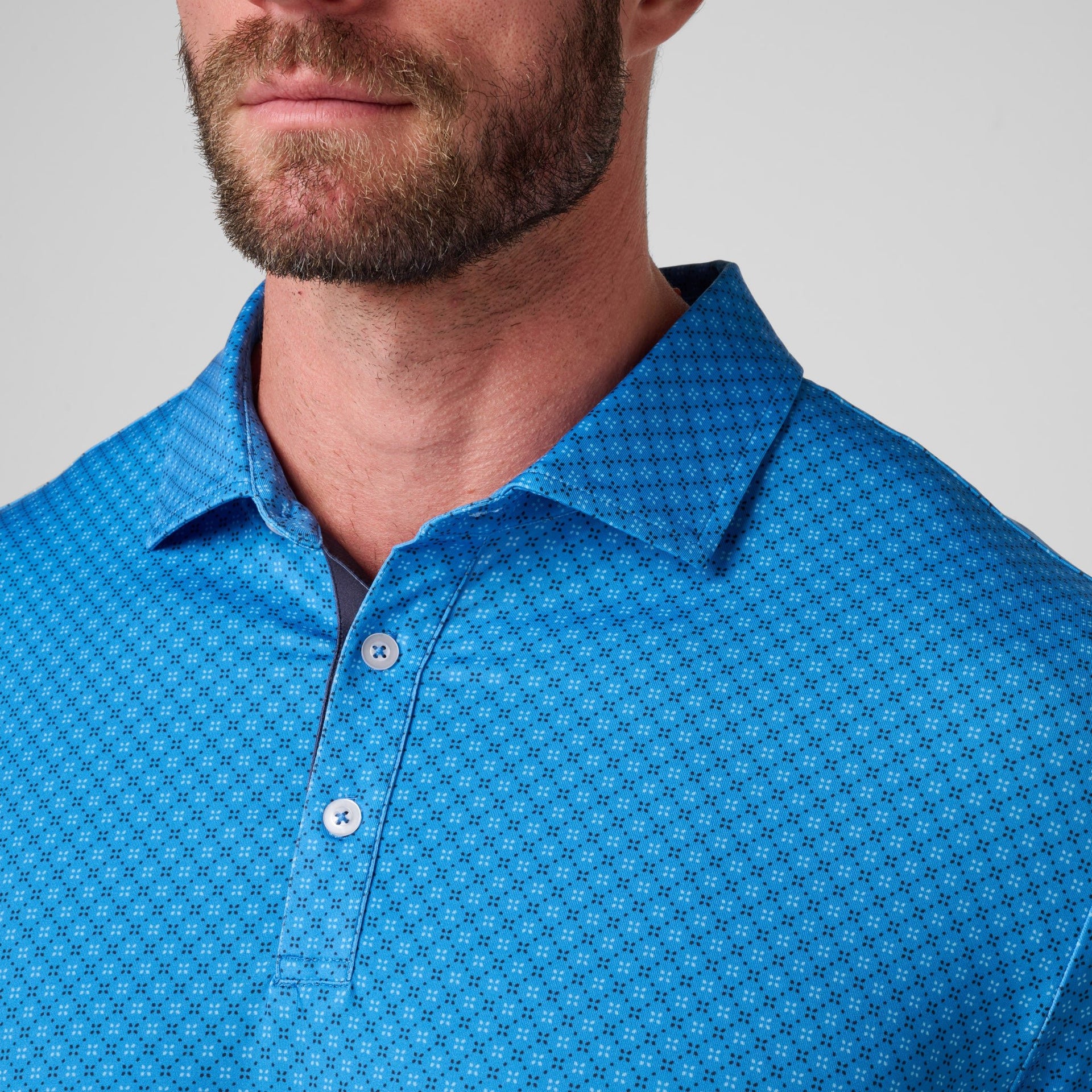 Bright blue polo shirt with small geometric dot print pattern and coral buttons on the chest placket.