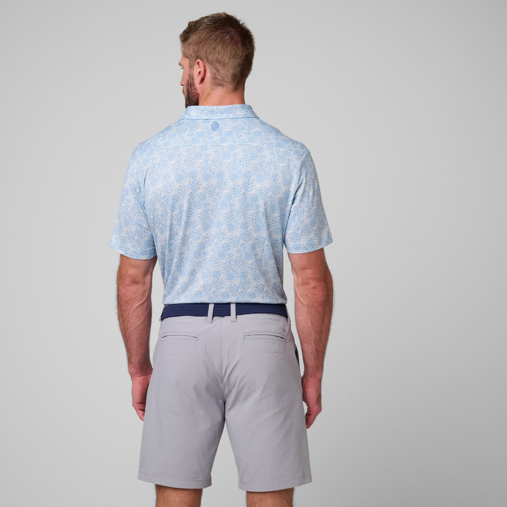 Man wearing light blue printed short-sleeve polo shirt and gray shorts with navy belt, shown from back view.