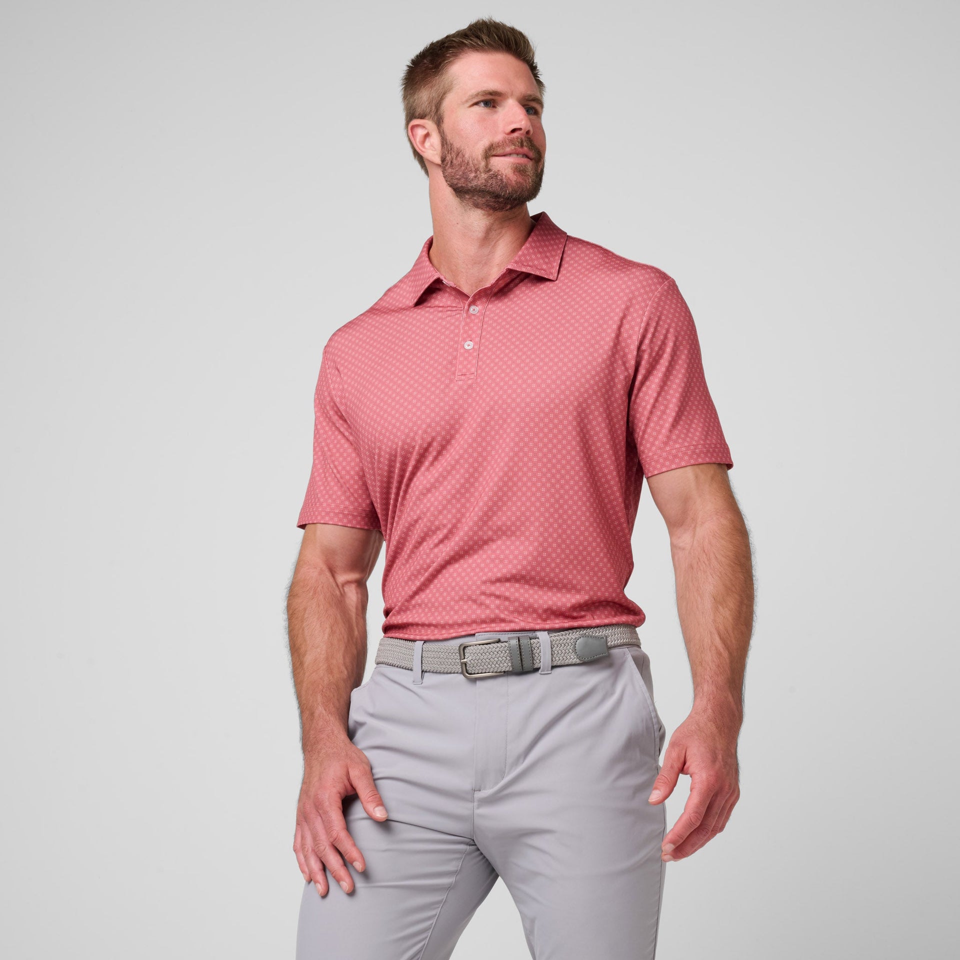 Man wearing rose pink polo with all-over diamond print pattern and light gray pants with gray belt.