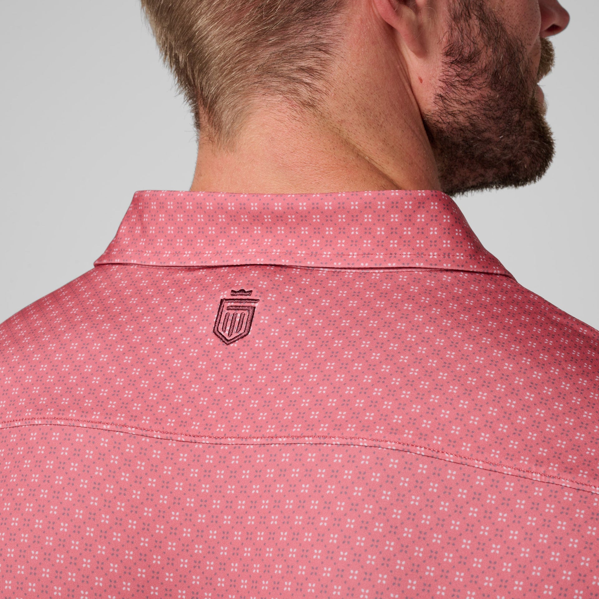 Man wearing pink polo with small geometric pattern and embroidered shield logo on back.