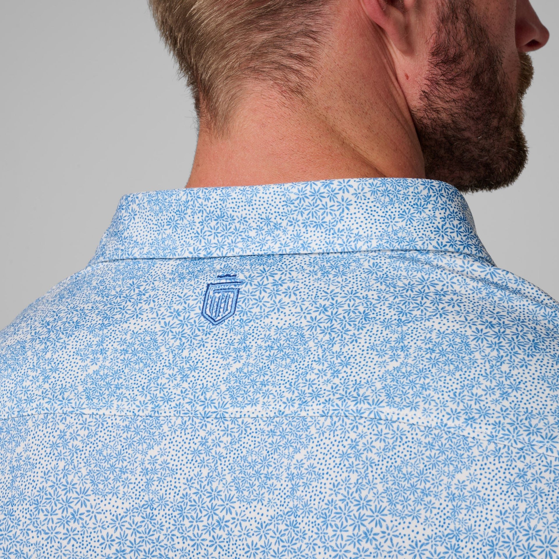 Back view of man wearing blue and white floral printed polo shirt with collar and embroidered logo.