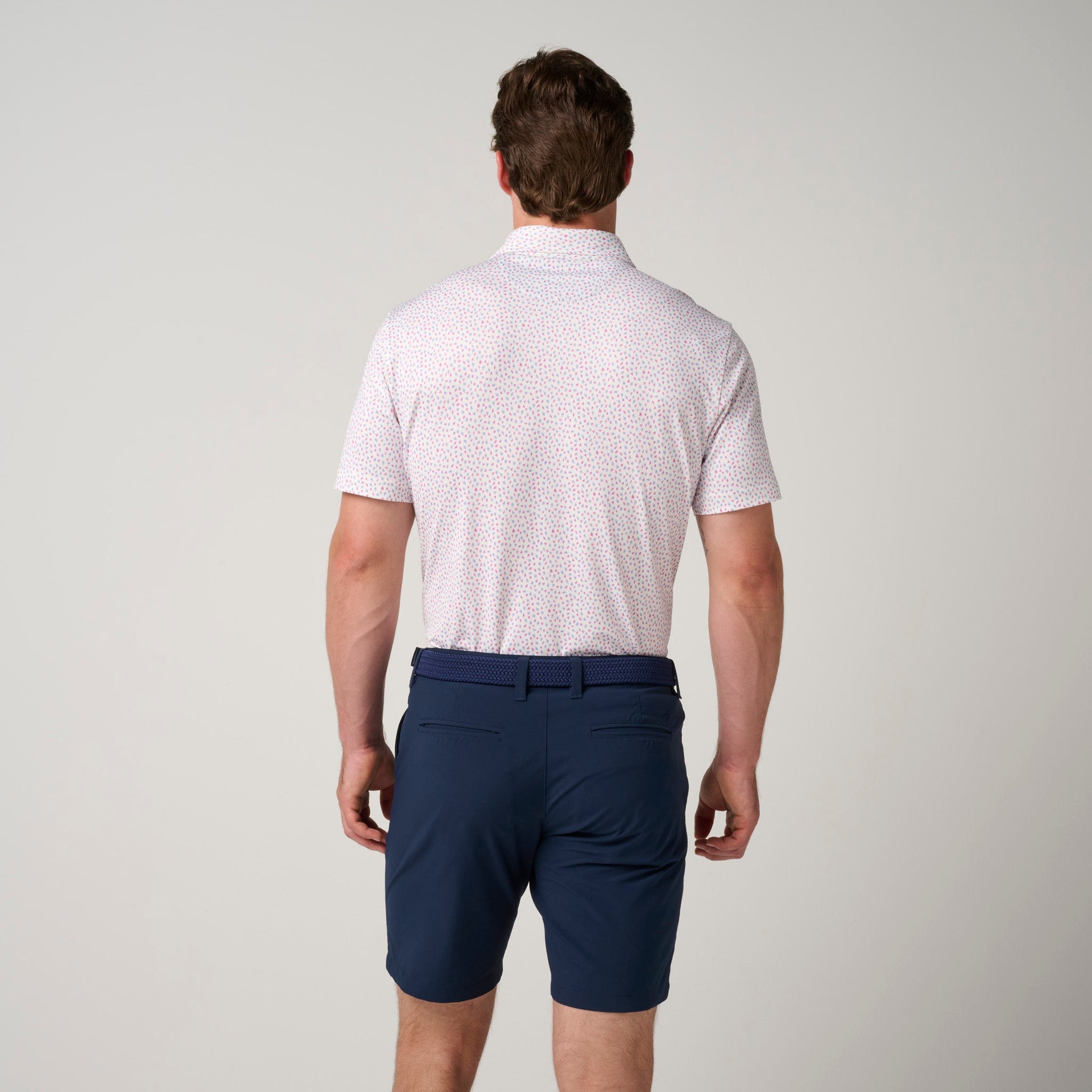 Man wearing light pink short-sleeve polo with small red pattern and navy shorts, shown from back.