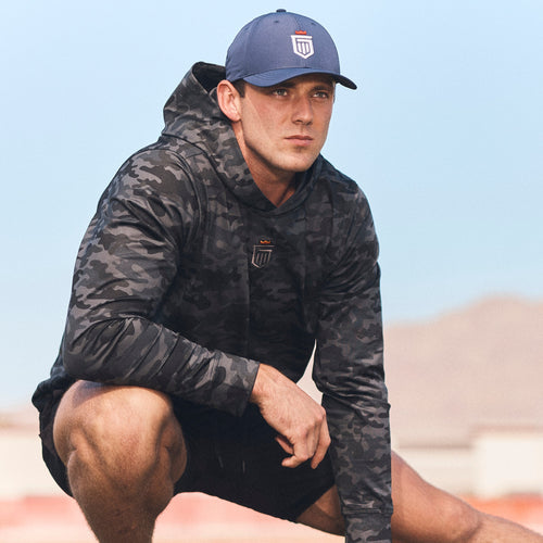 Man wearing black camouflage hoodie with small shield logo on chest, paired with blue baseball cap.