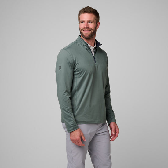 Man wearing sage green quarter zip pullover with dark collar and small chest logo over gray pants.
