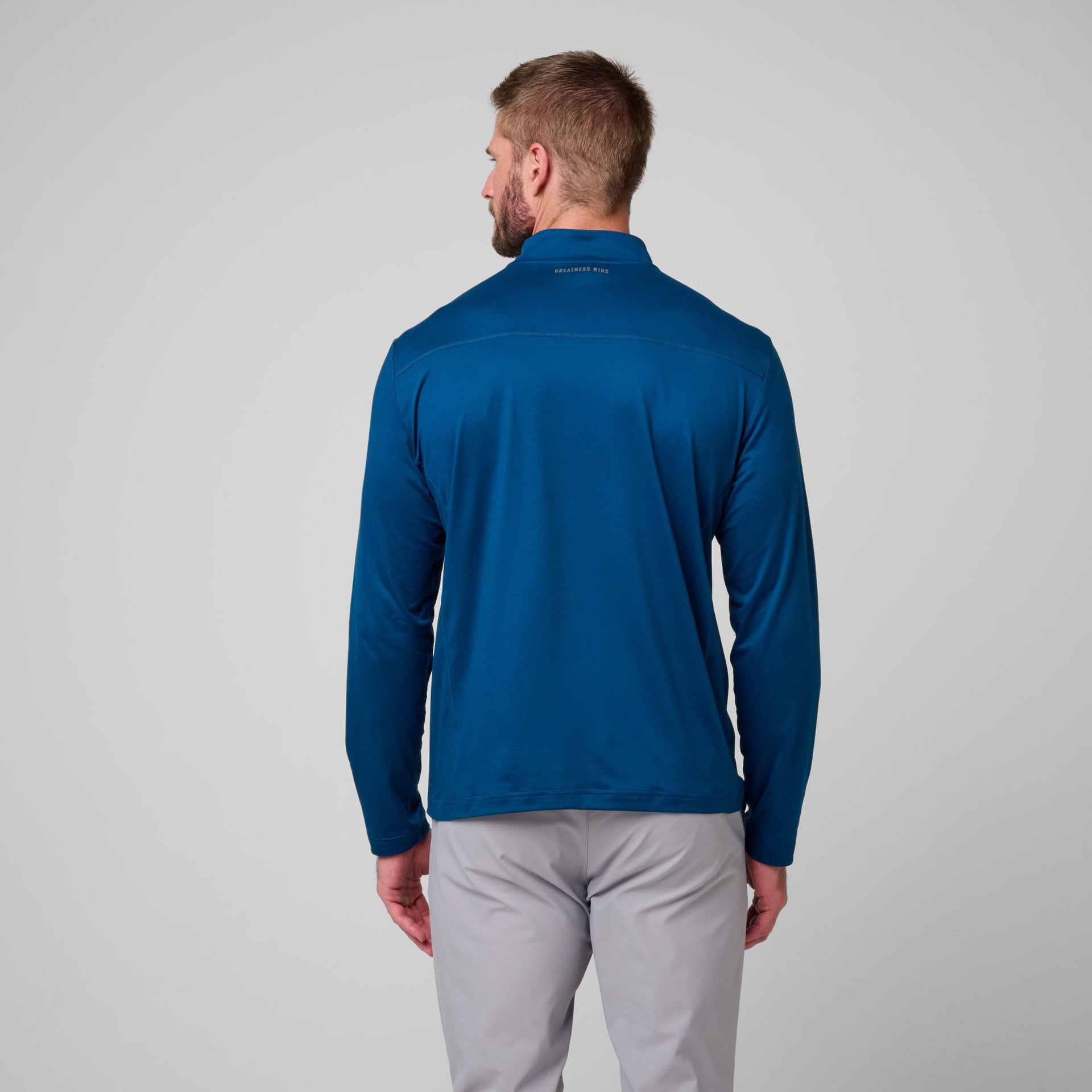 Man wearing blue quarter zip pullover with long sleeves and gray pants, shown from back view.