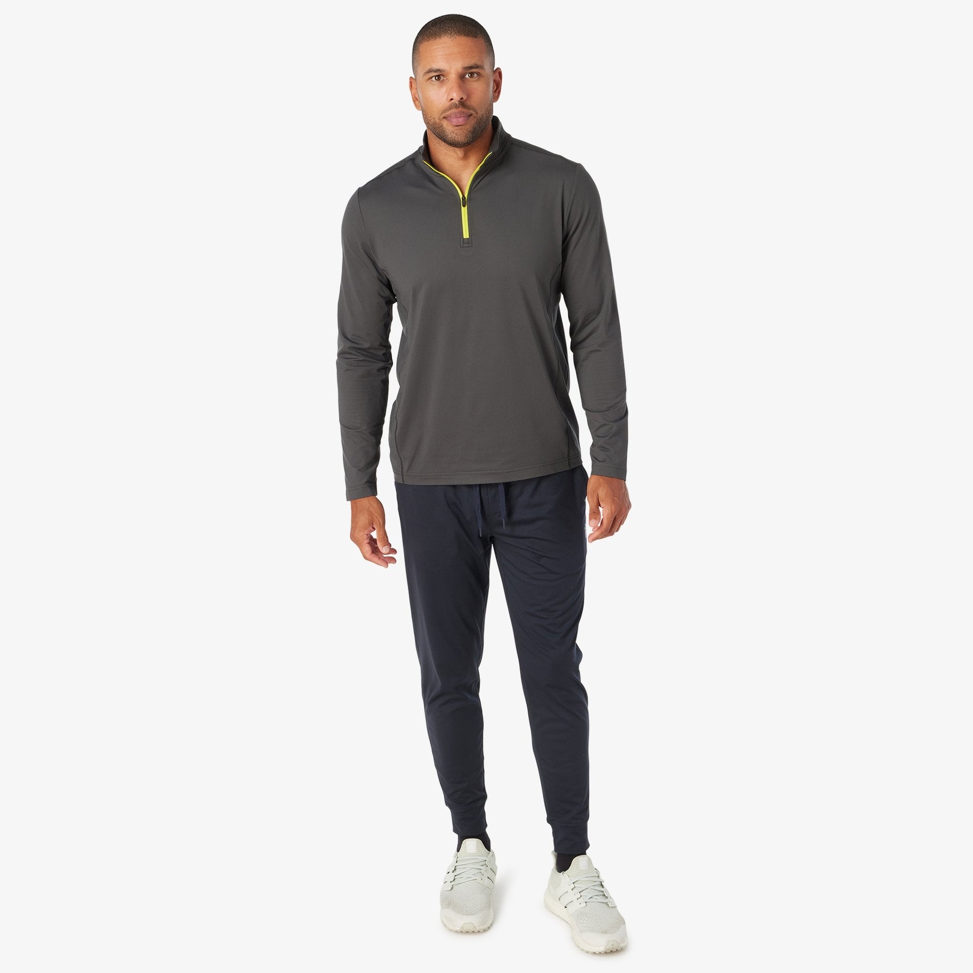 Man wearing charcoal quarter-zip pullover with yellow zipper and navy joggers with white sneakers.