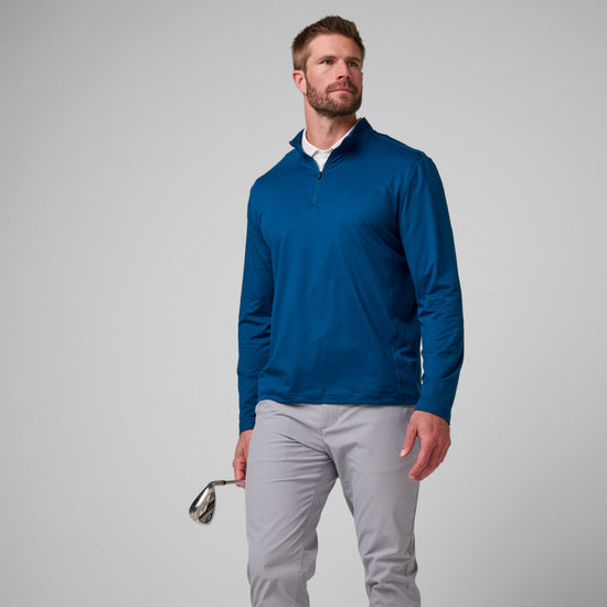 Man wearing a navy blue quarter-zip pullover with long sleeves and gray pants, holding golf shoes.