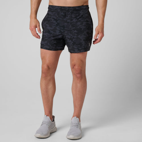 Black camouflage athletic shorts with elastic waistband on male model wearing gray sneakers.