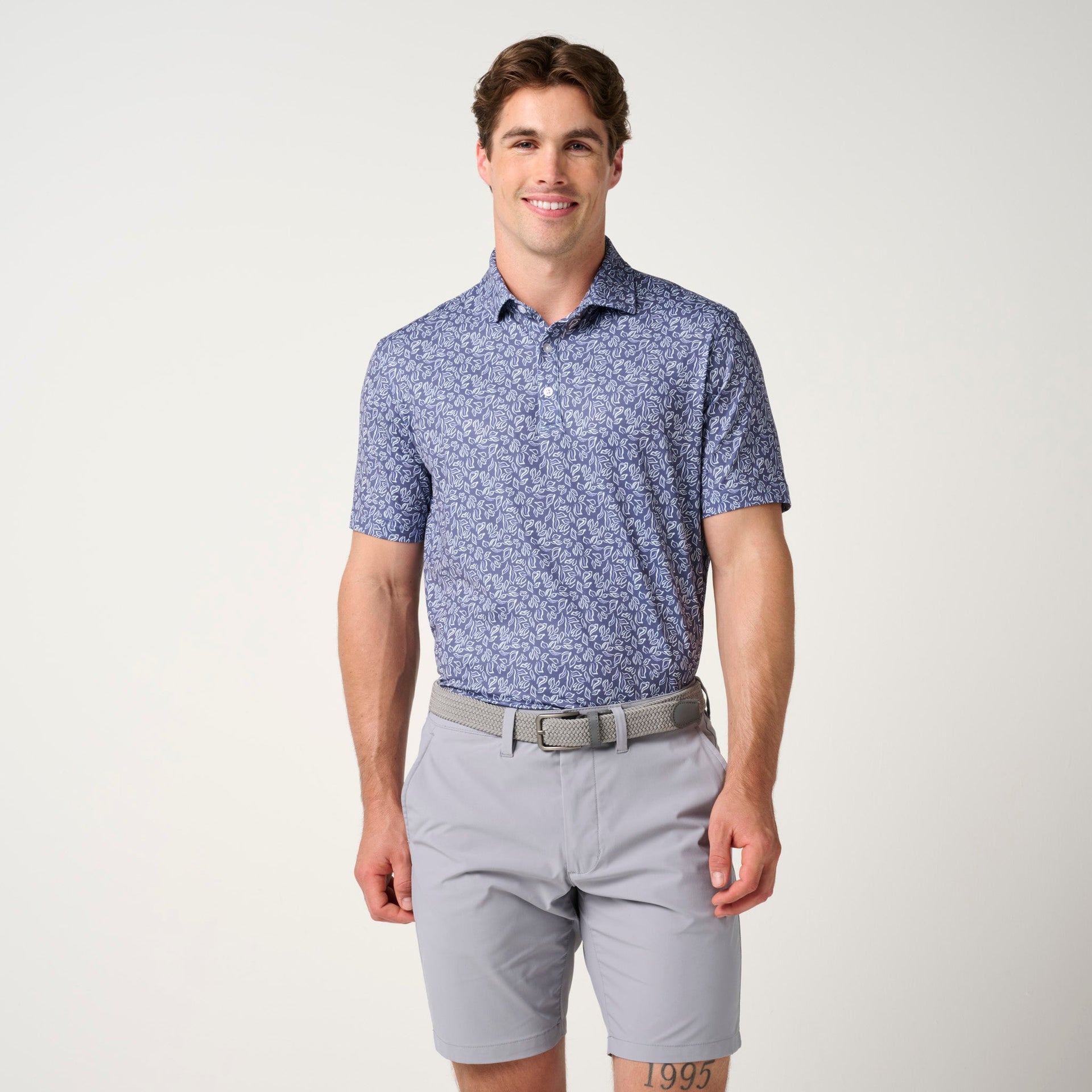 Man wearing blue floral polo shirt with gray shorts and tan belt against neutral background.