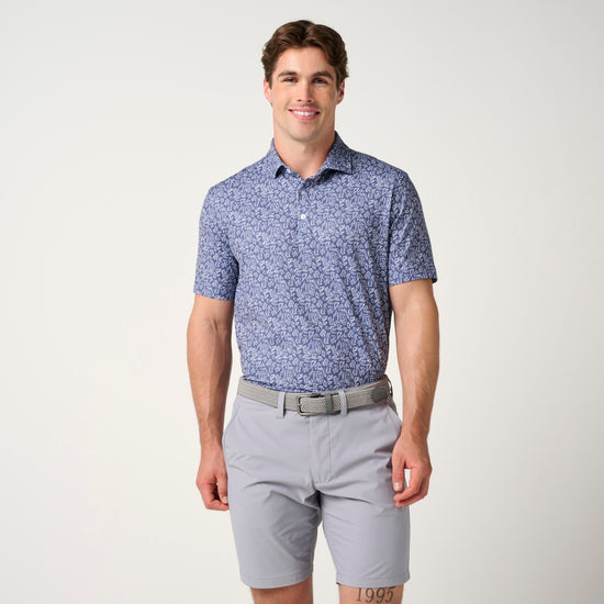 Man wearing blue floral polo shirt with gray shorts and tan belt against neutral background.