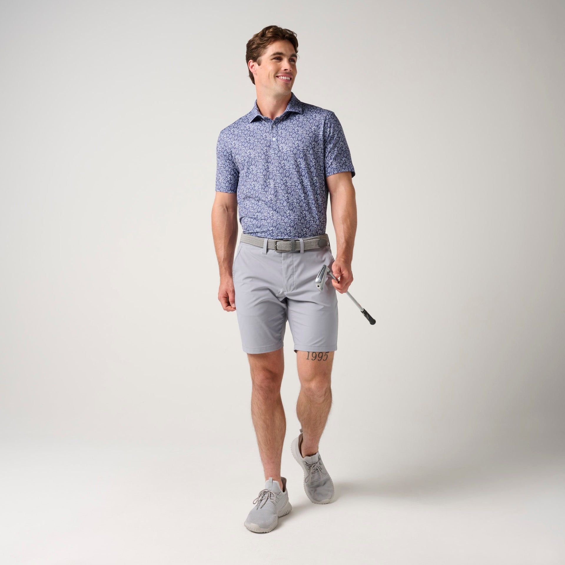 Man wearing blue floral polo shirt with gray shorts and tan belt, holding a golf club.