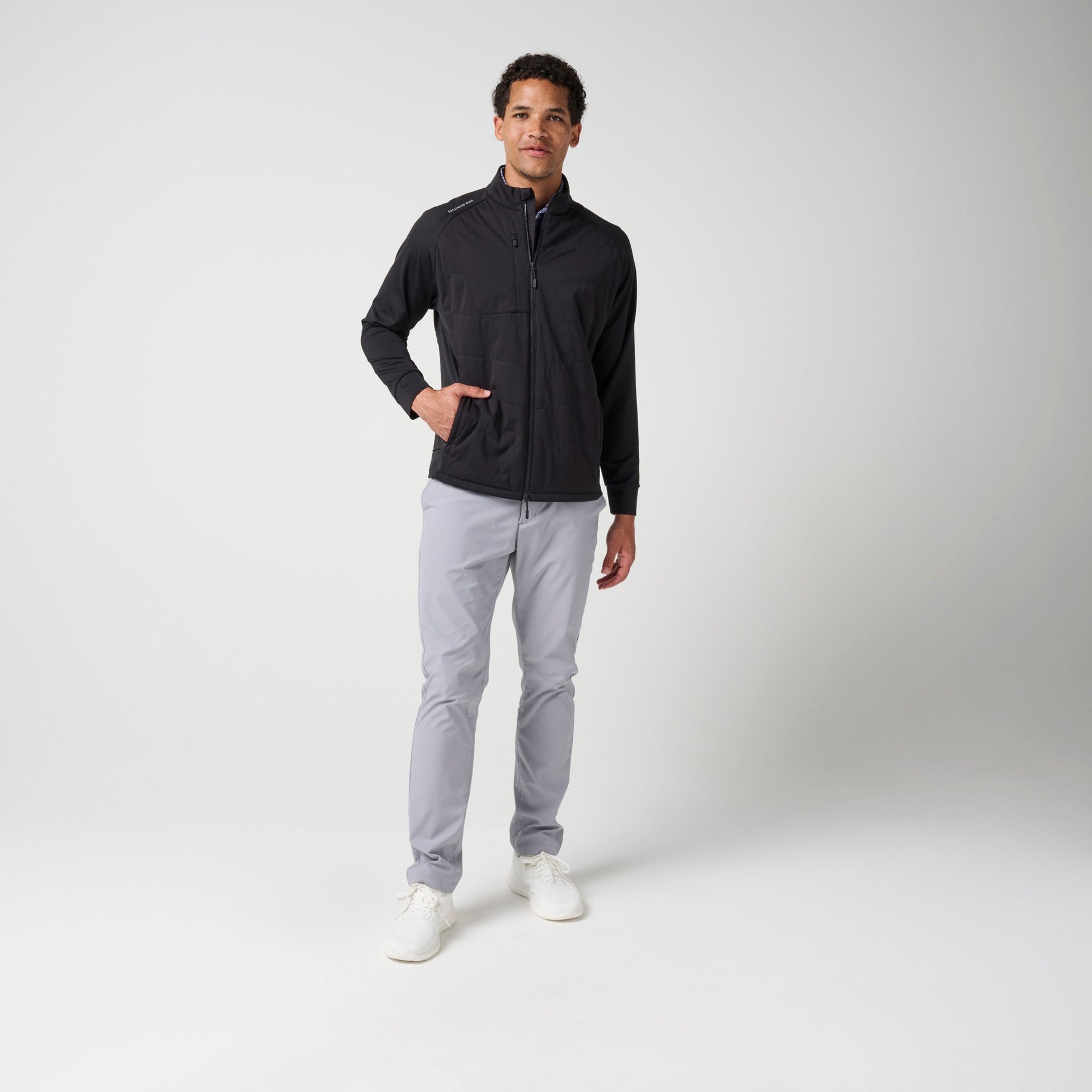 Black full-zip hybrid jacket with ribbed sleeves worn with light gray pants.