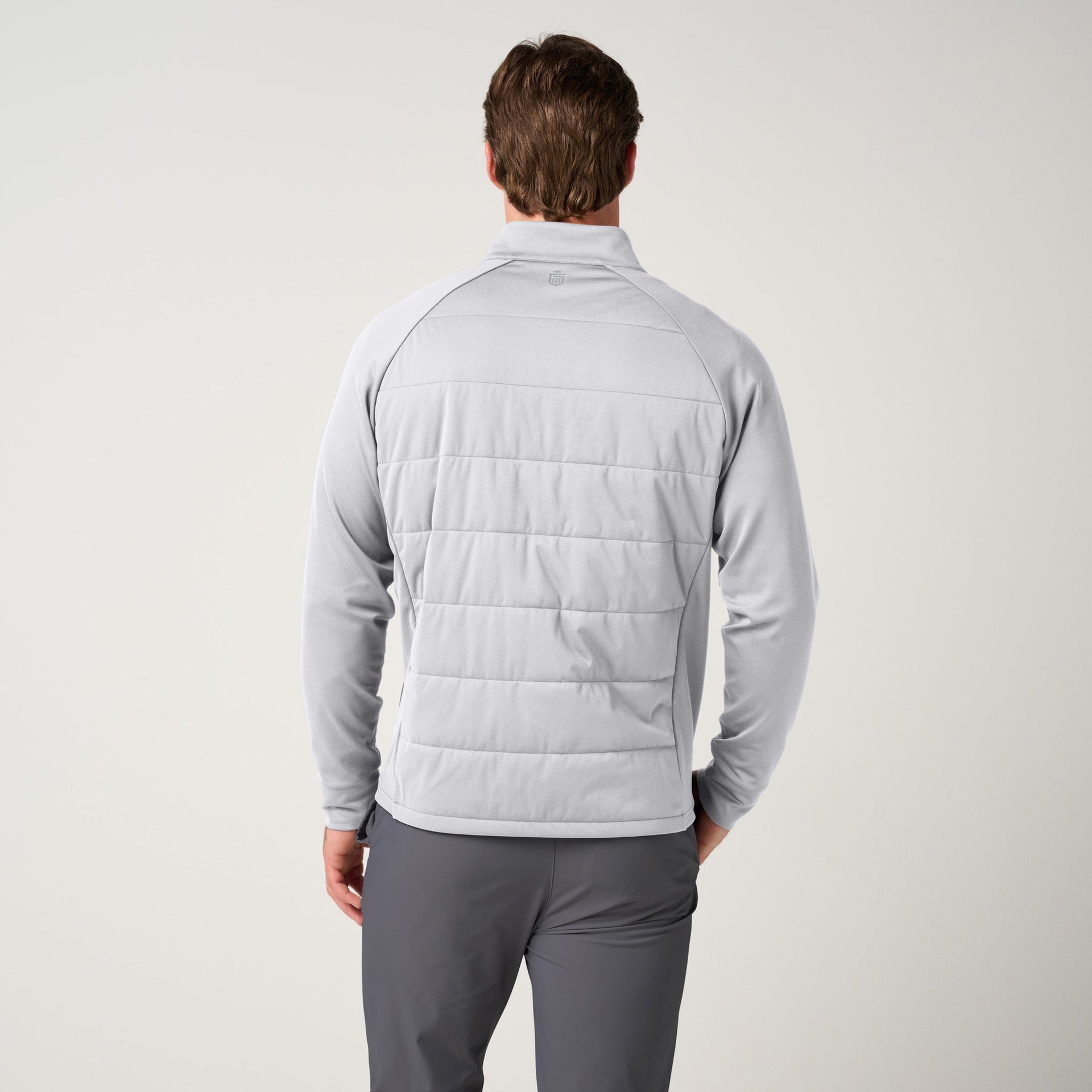 Man wearing light gray hybrid jacket with quilted back panel and knit sleeves, paired with charcoal pants, shown from behind.