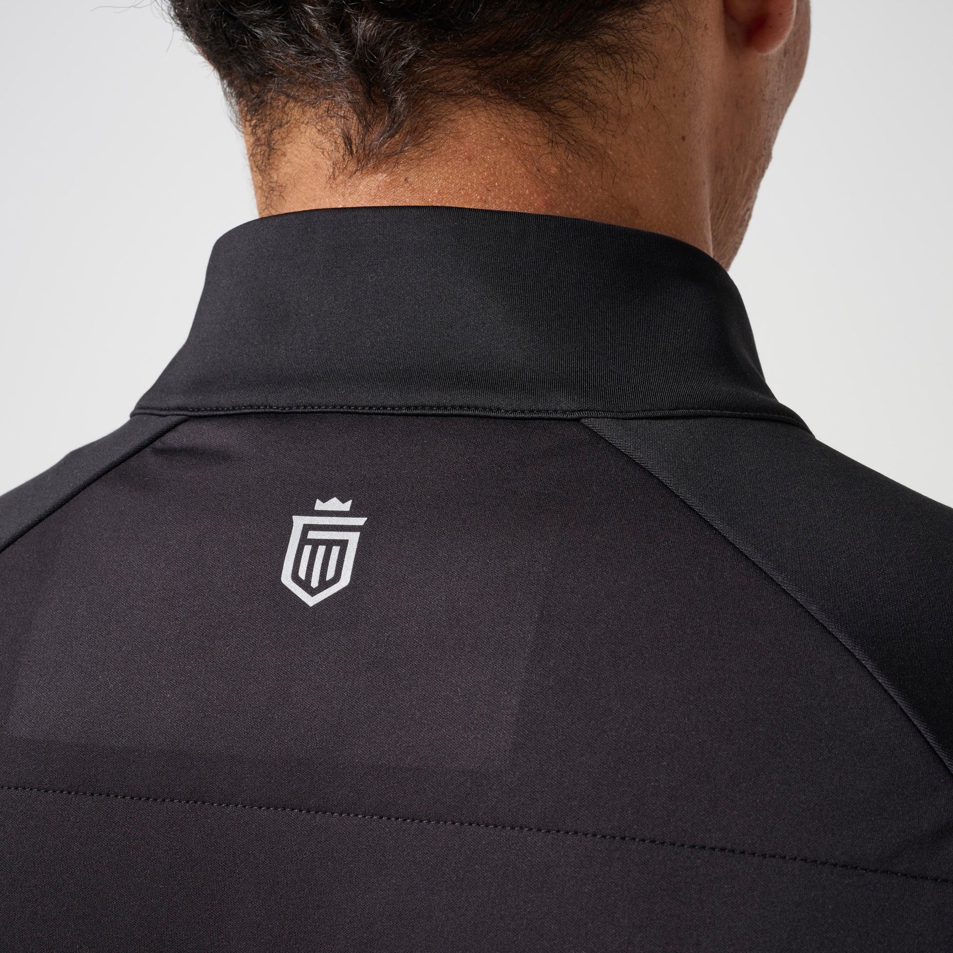 Black hybrid jacket with ribbed collar and white shield logo on back, shown on male model from neck down.