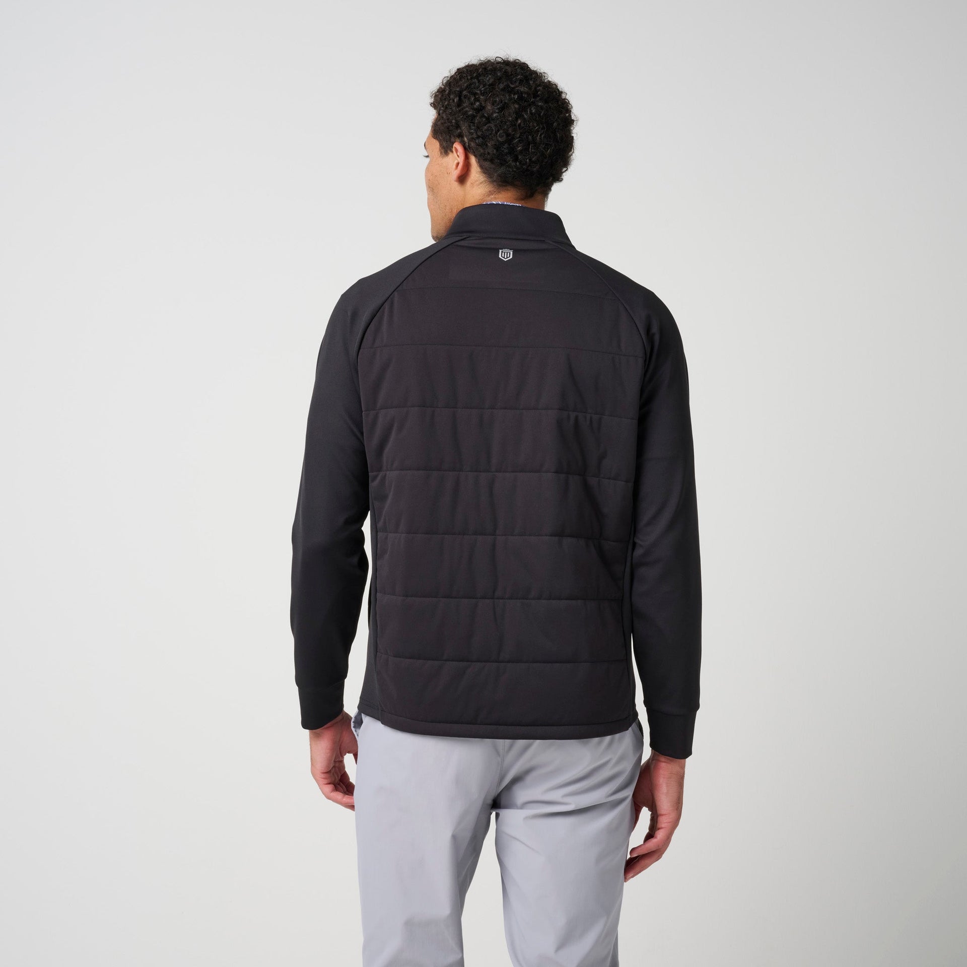 Black hybrid jacket with quilted back panel and long sleeves worn over light gray pants, shown from back view.