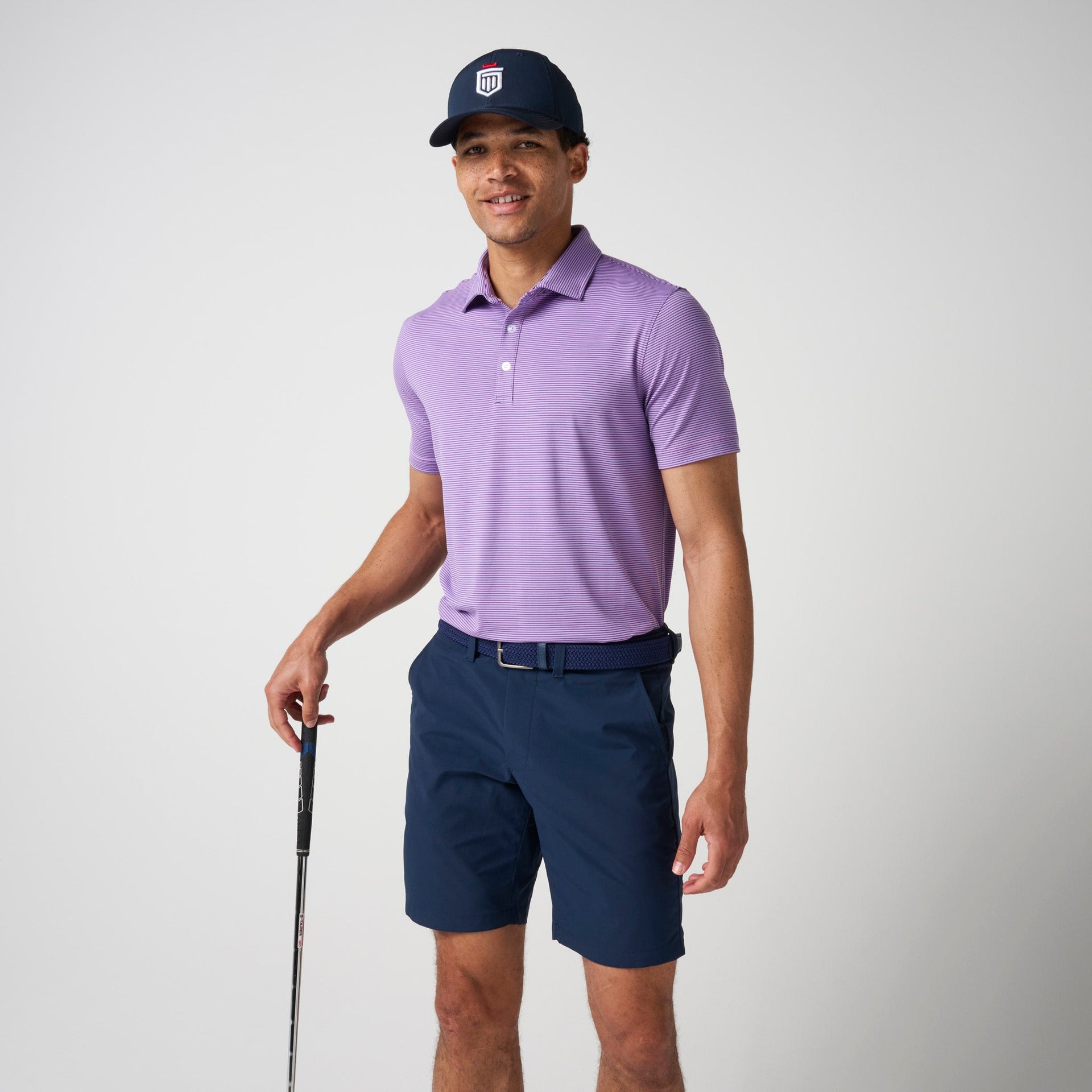 Man wearing lavender striped polo shirt with navy shorts and dark cap, holding a golf club.