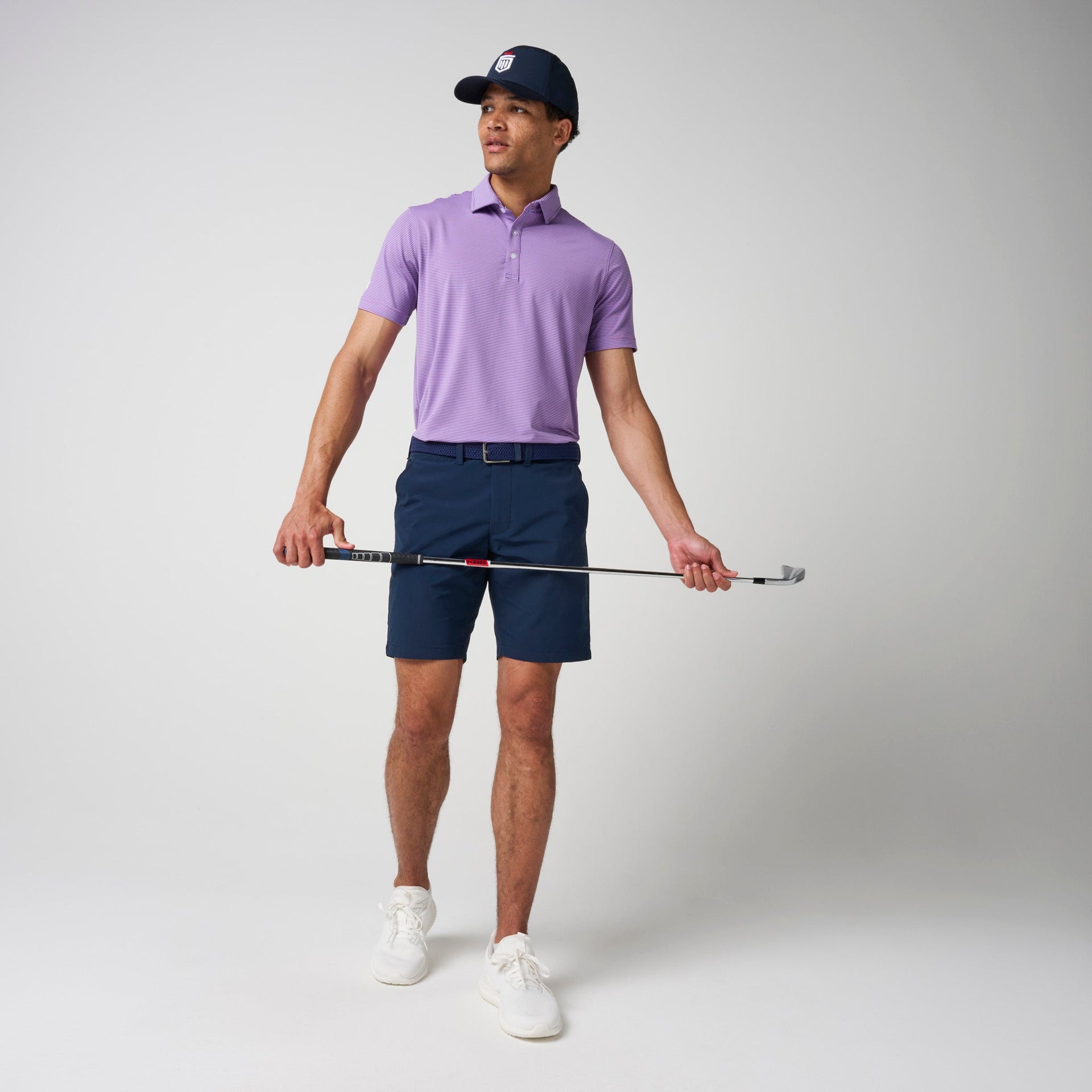 Man wearing lavender polo shirt with navy shorts, belt, and cap, holding a golf club.